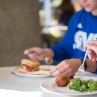 Three plates of food, one with broccoli and a chicken wing, and a hamburger. Two students are seen in the photo.